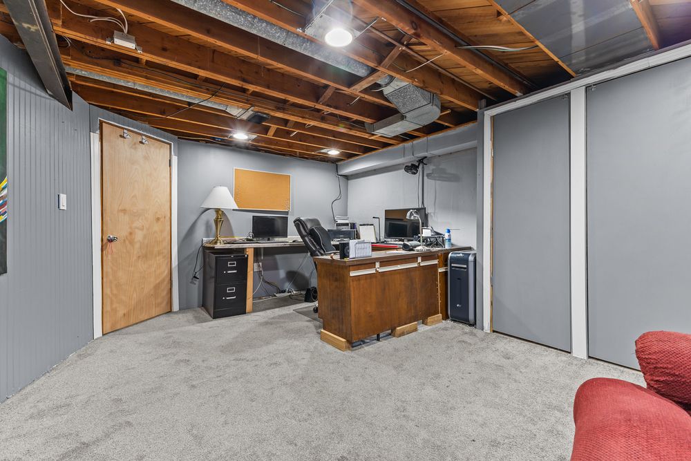 Finished basement office space with gray carpet, exposed ceiling, wooden desk, and computer workstation.