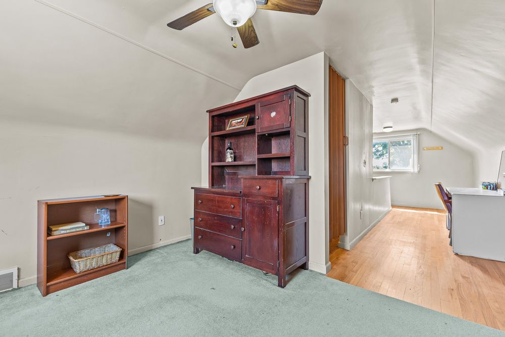 Carpeted attic room with vaulted ceiling, antique dresser, ceiling fan, and wood floors.