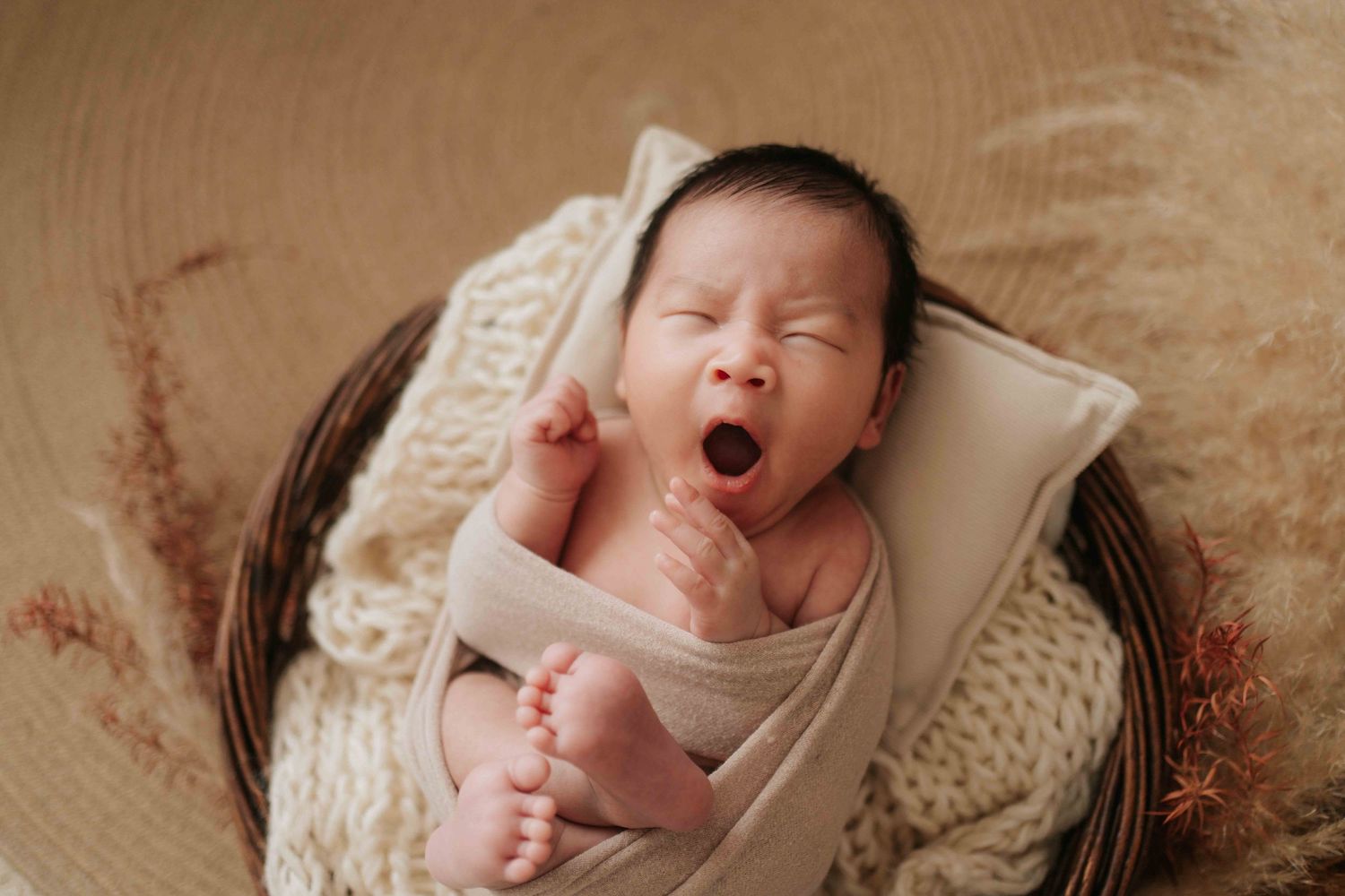 Newborn baby yawning while swaddled in a cream knit wrap, resting in a wicker basket for a studio photo.