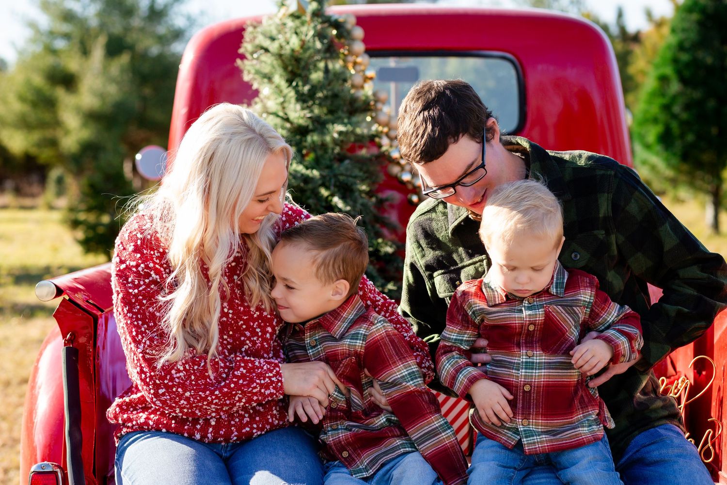 Family of four in matching plaid shirts sitting on red truck tailgate at Christmas tree farm.
