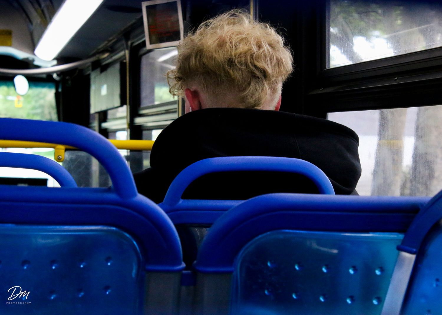 Blonde person sitting alone on blue seats inside a public bus.