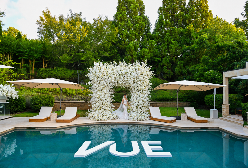 Bride poses under white floral arch by pool with VUE letters, lounge chairs, and umbrellas at outdoor wedding venue.