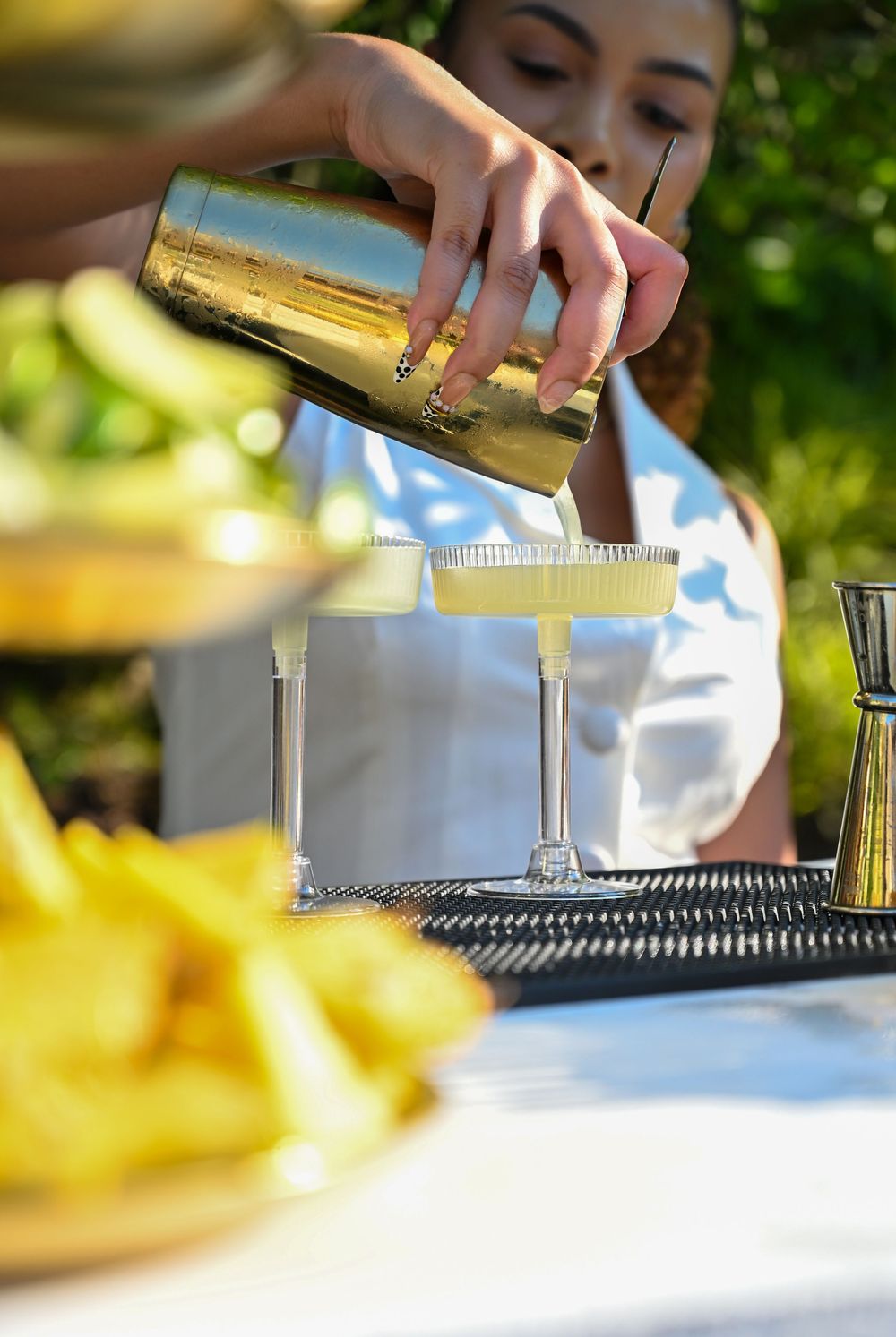 Woman pouring yellow cocktail from gold shaker into coupe glasses on outdoor bar tray with fruit garnishes.