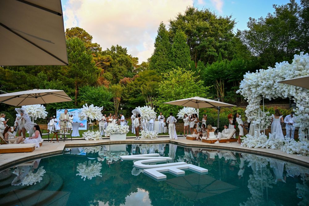 Elegant all-white poolside wedding reception with floral arches, white lounge floats, and lush green trees at dusk.