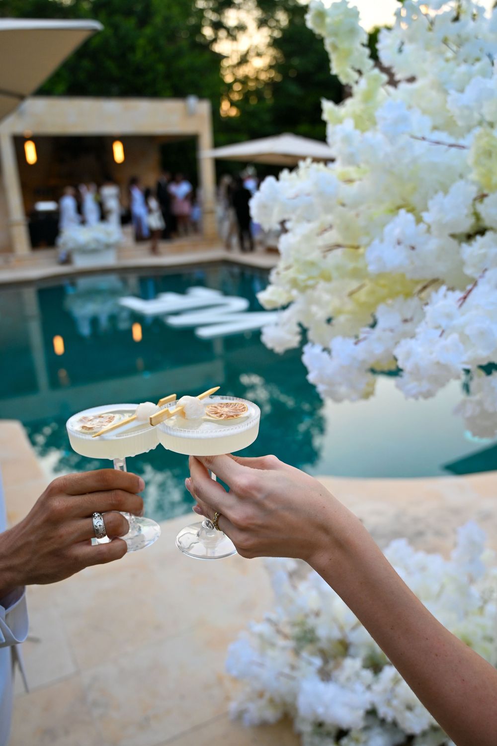 Two hands clinking white cocktails garnished with flowers near a pool at an elegant outdoor wedding reception.