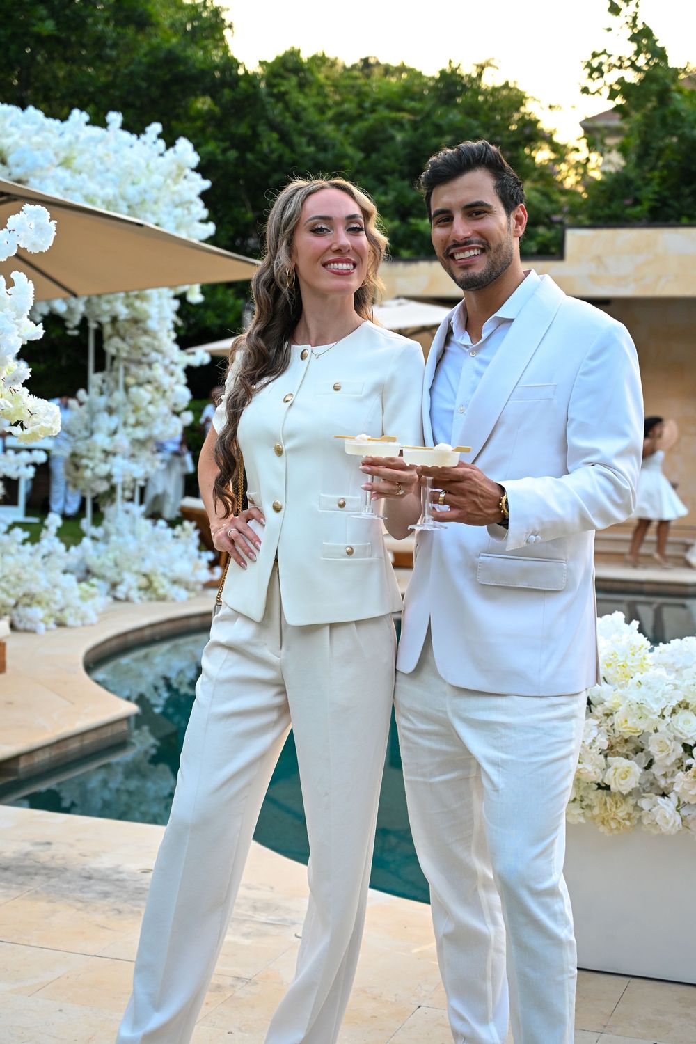 Elegant couple in white suits toasting with cocktails at a luxurious poolside wedding event with floral decor.