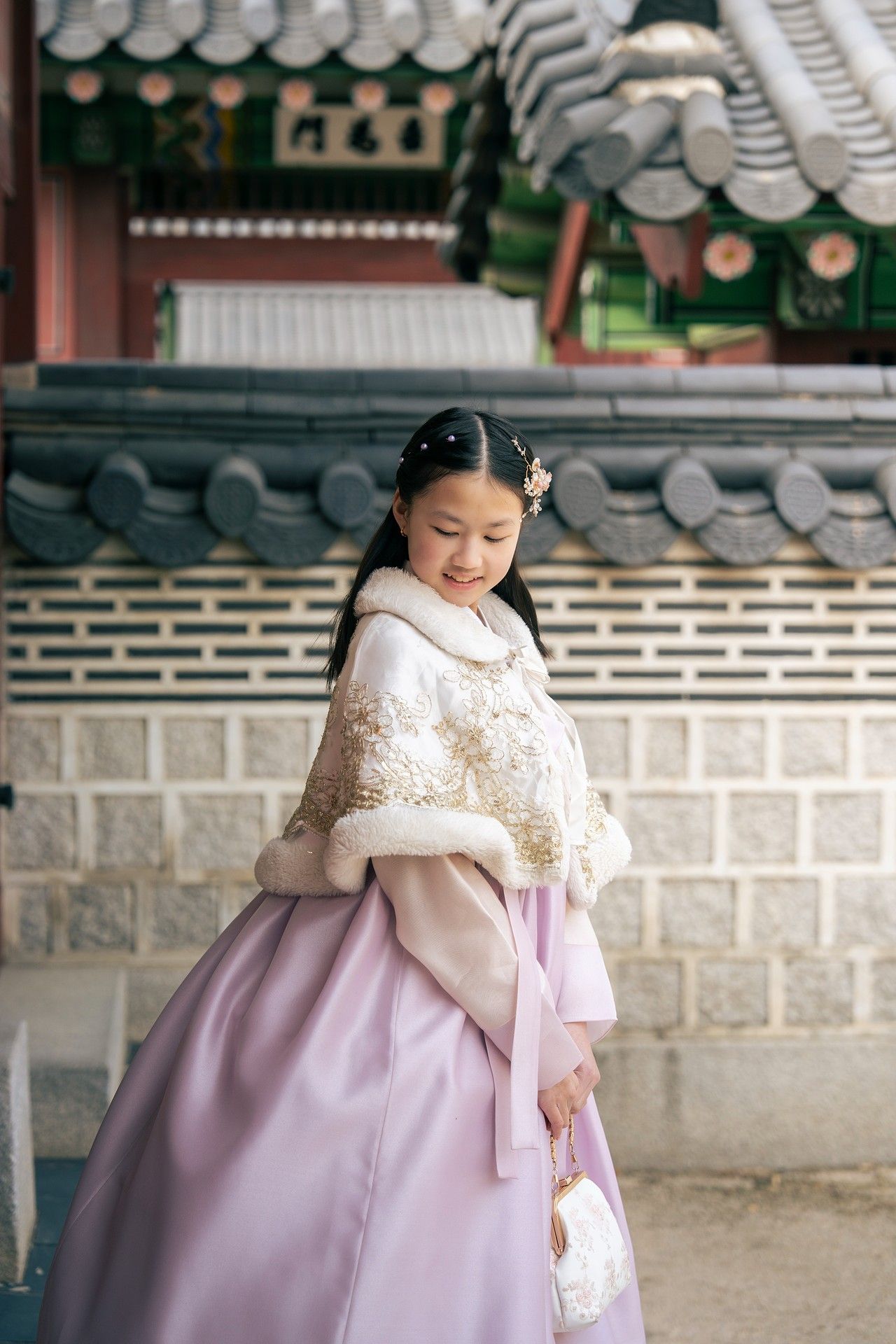 Young girl in traditional Korean hanbok with white fur cape smiles at historic palace courtyard.