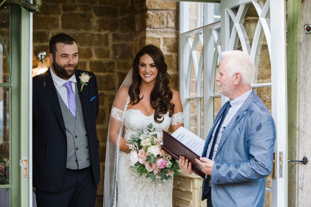 Bride and groom stand with officiant during outdoor wedding ceremony, bride holding pink and white bouquet in elegant lace gown.