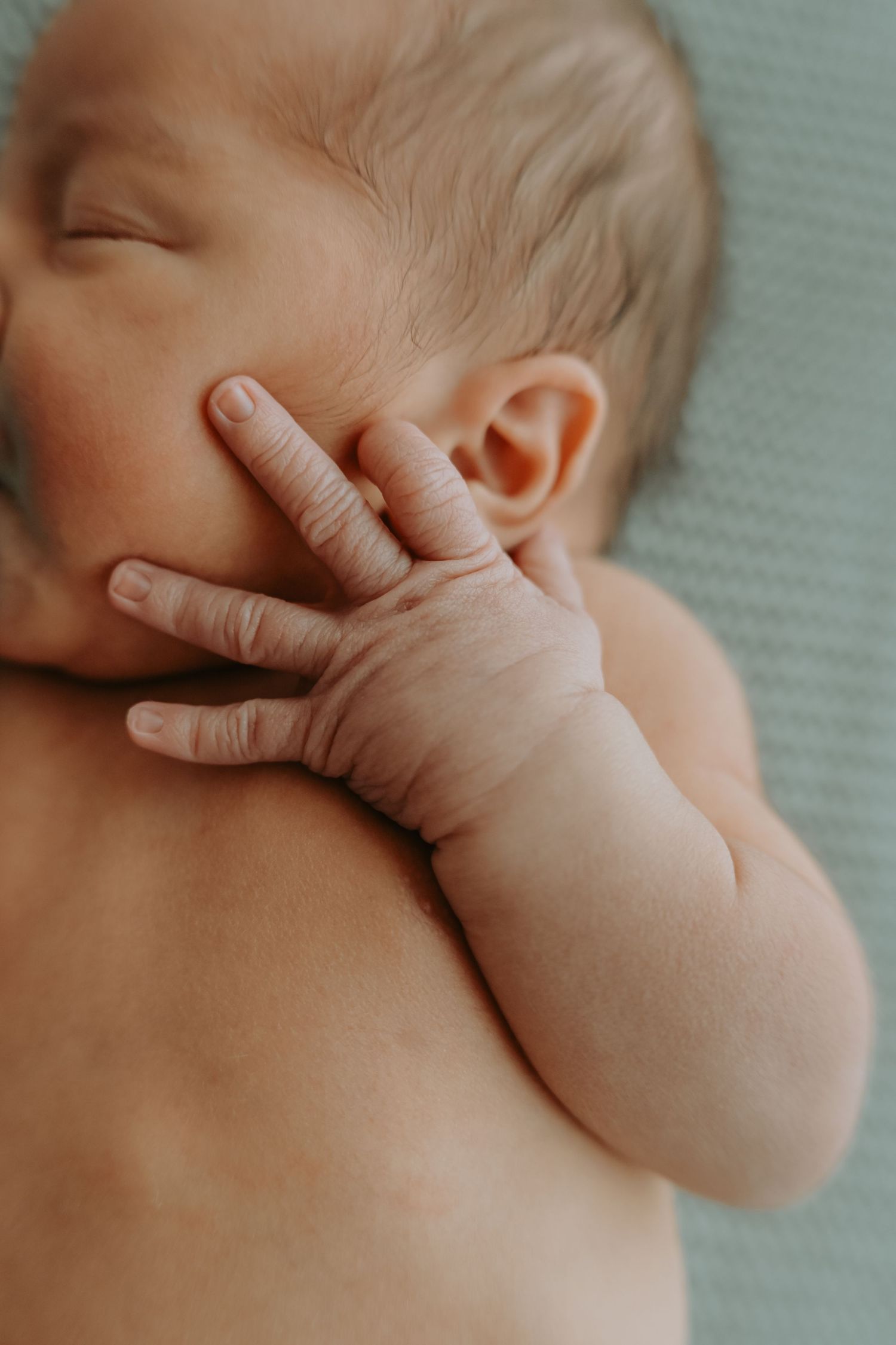Close-up of a sleeping newborn baby's tiny hand resting against their cheek, lying on a soft light blue blanket.