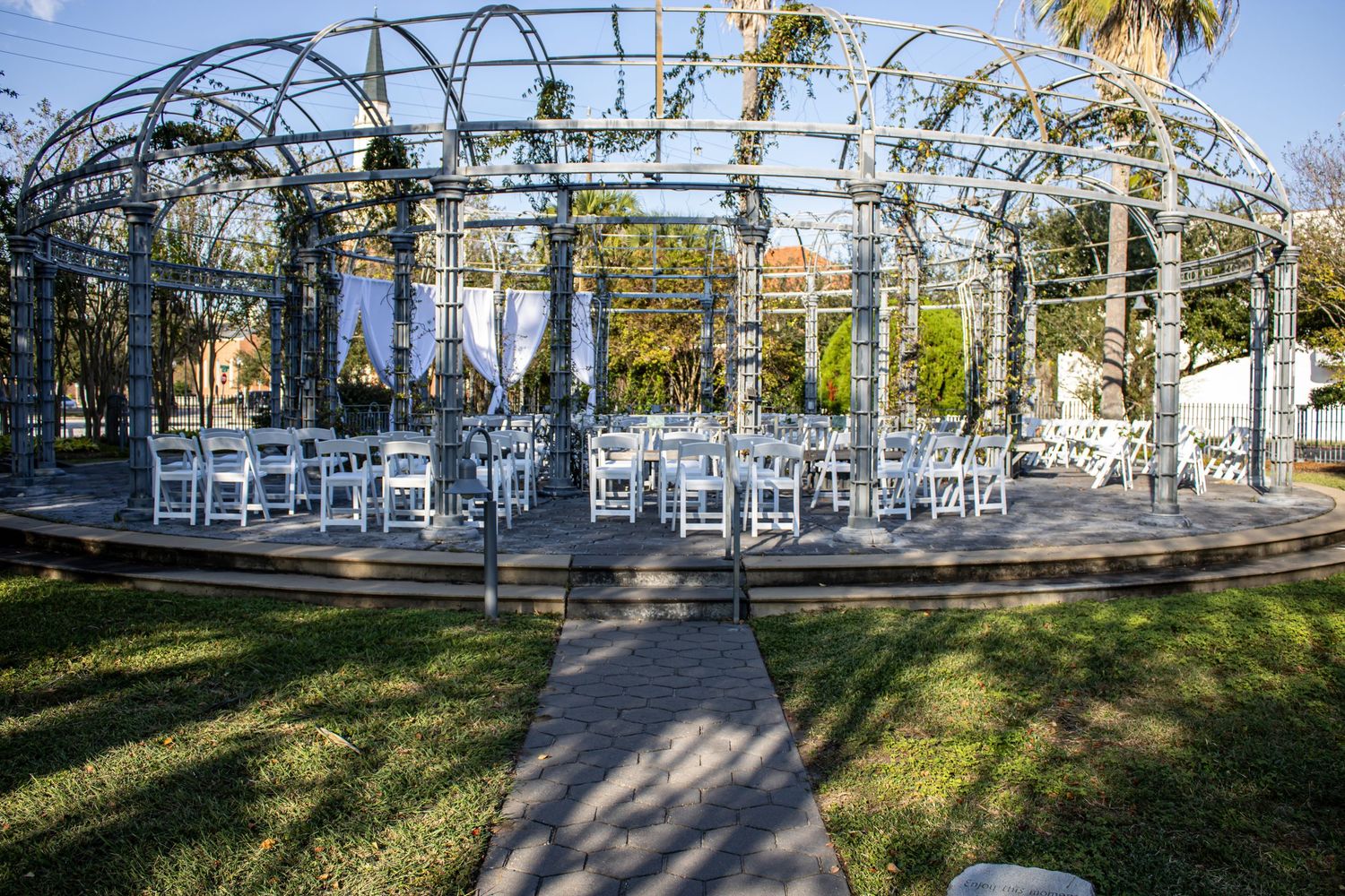 Elegant outdoor wedding venue with white chairs under a metal pergola arch surrounded by lush greenery and draped fabric.