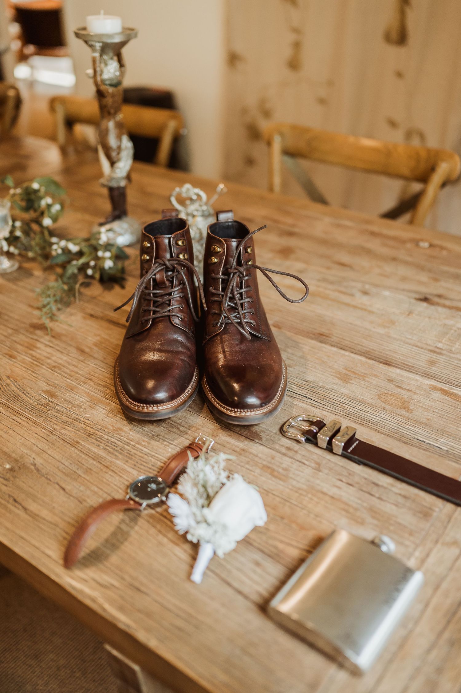 Brown leather boots with groom accessories including a watch, boutonniere, belt, and flask on a rustic wooden table.