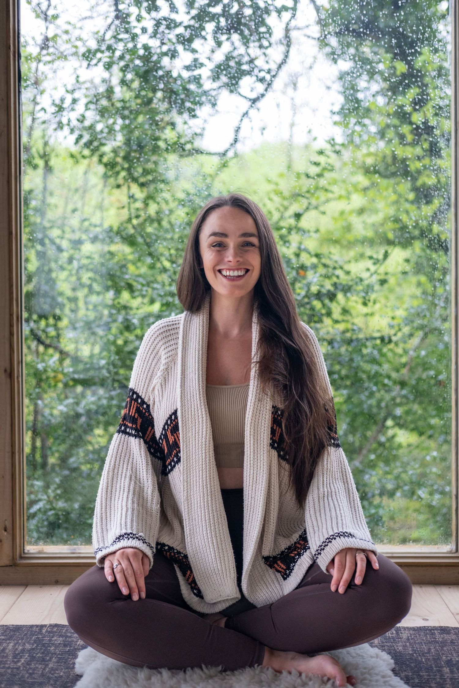 Smiling young woman sitting cross-legged on a fluffy rug in a cozy cardigan, meditating by a large window with green trees.