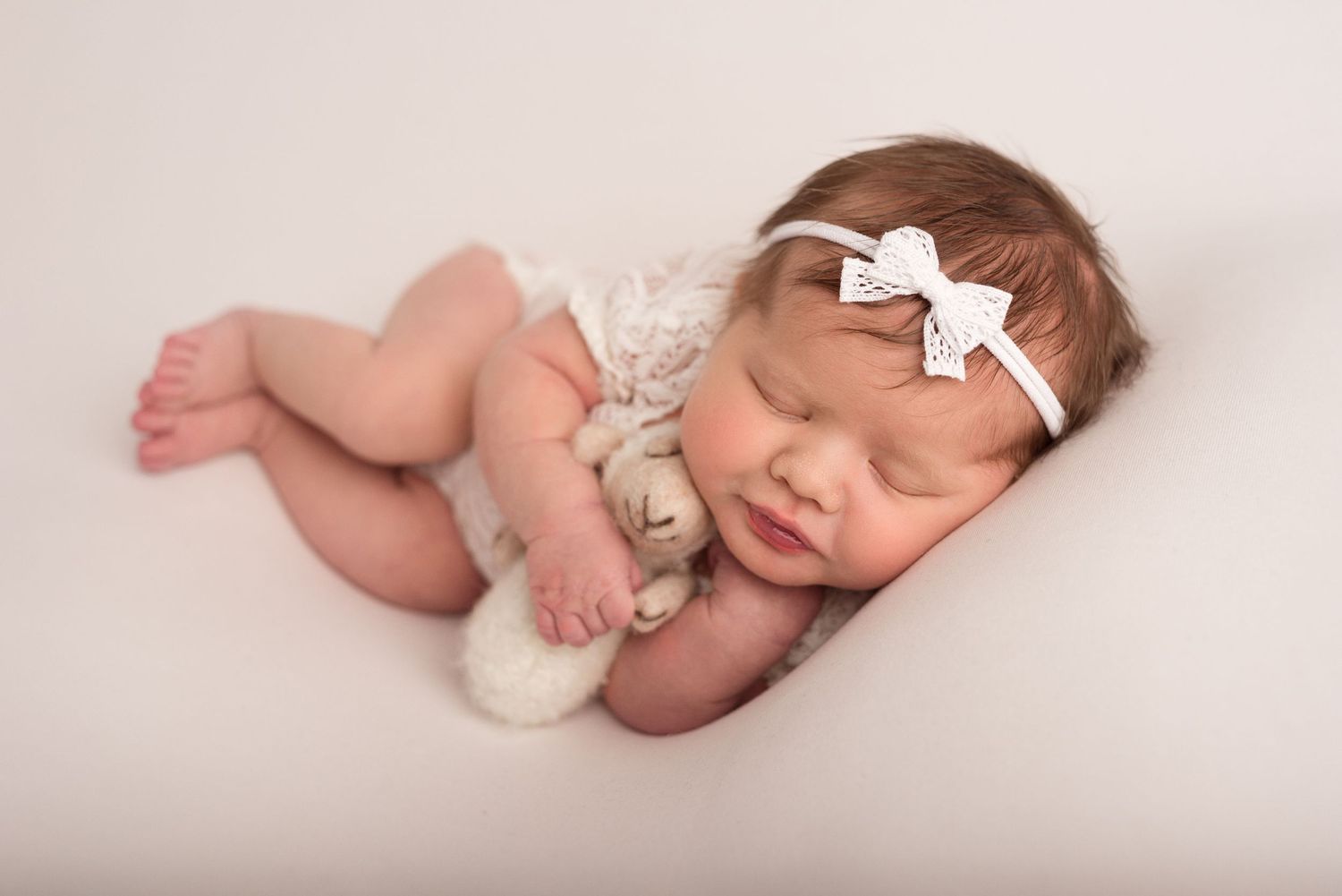 Sleeping newborn baby wearing a white knit bear bonnet hat, curled up peacefully on a soft white background.