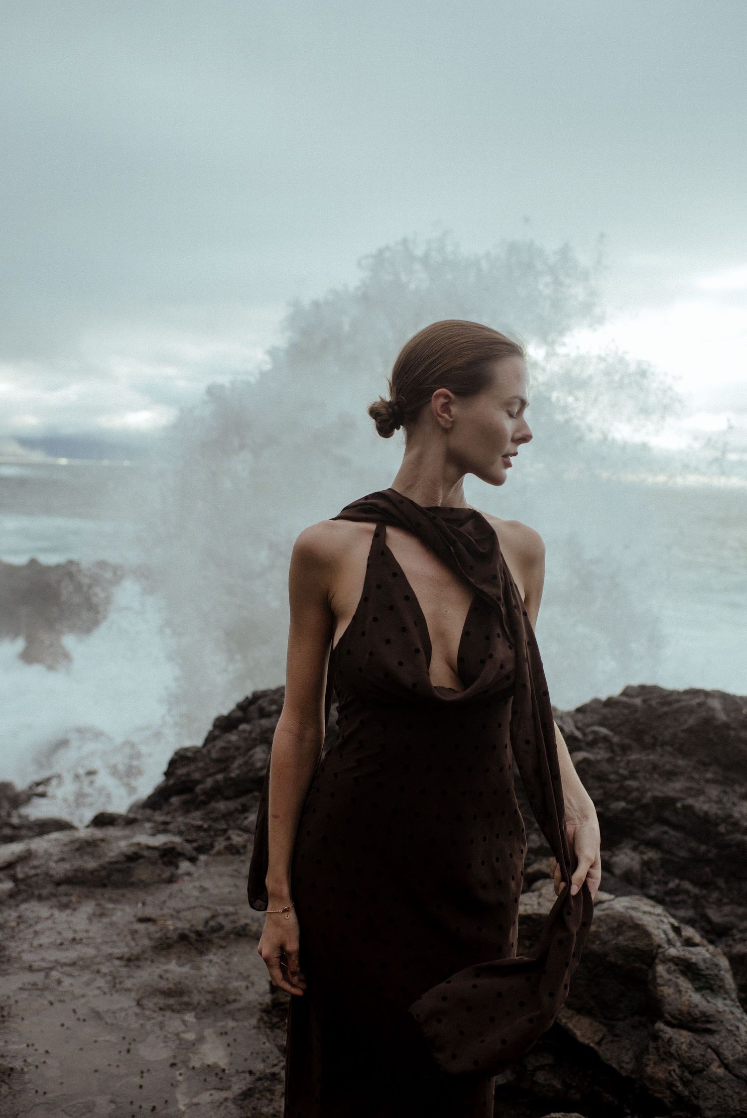 Woman in elegant black backless gown standing on rocky shore with dramatic ocean wave crashing behind her.