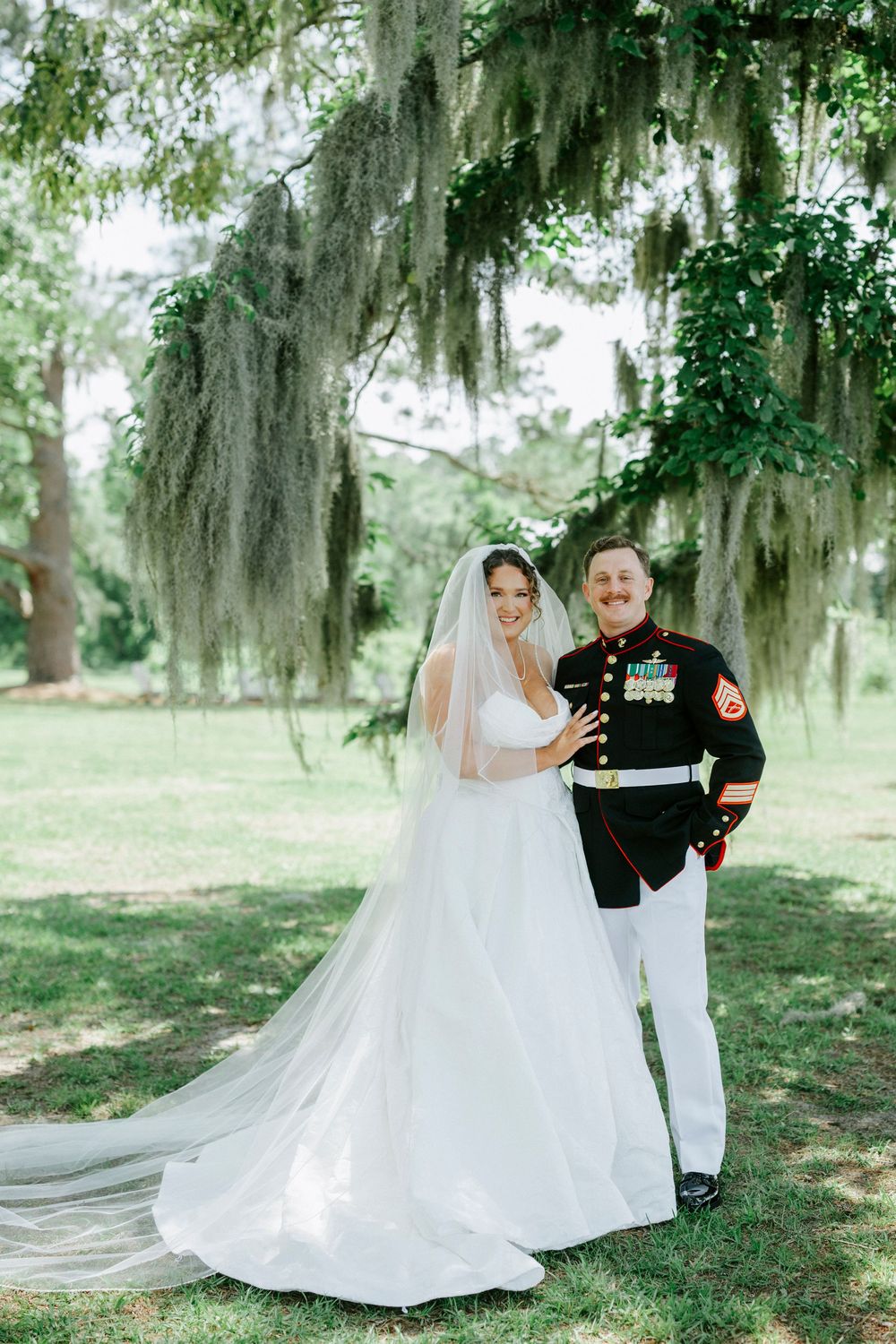 Bride in white gown and groom in Marine dress blues pose under Spanish moss-draped trees.