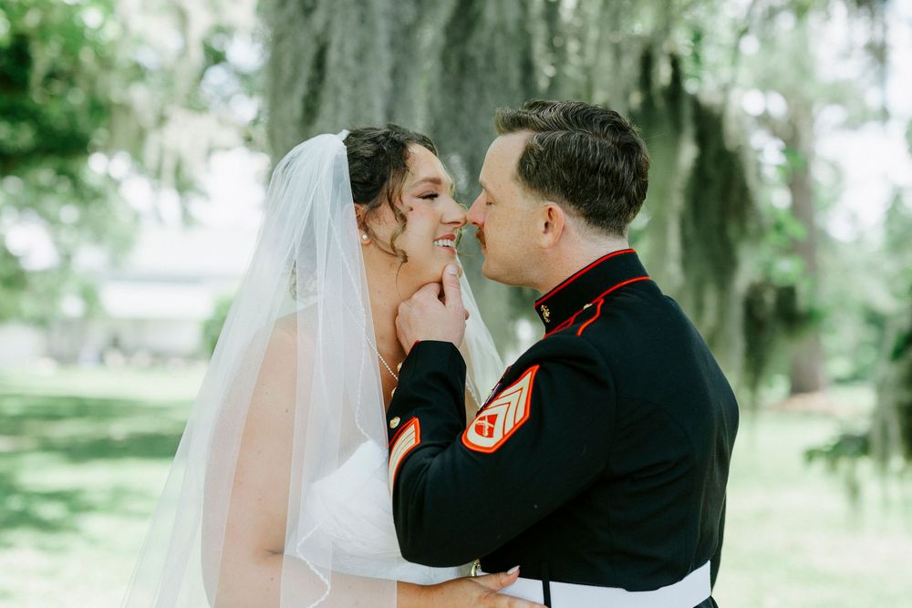 A bride in a white veil and groom in Marine dress uniform share a tender moment outdoors under moss-draped trees.