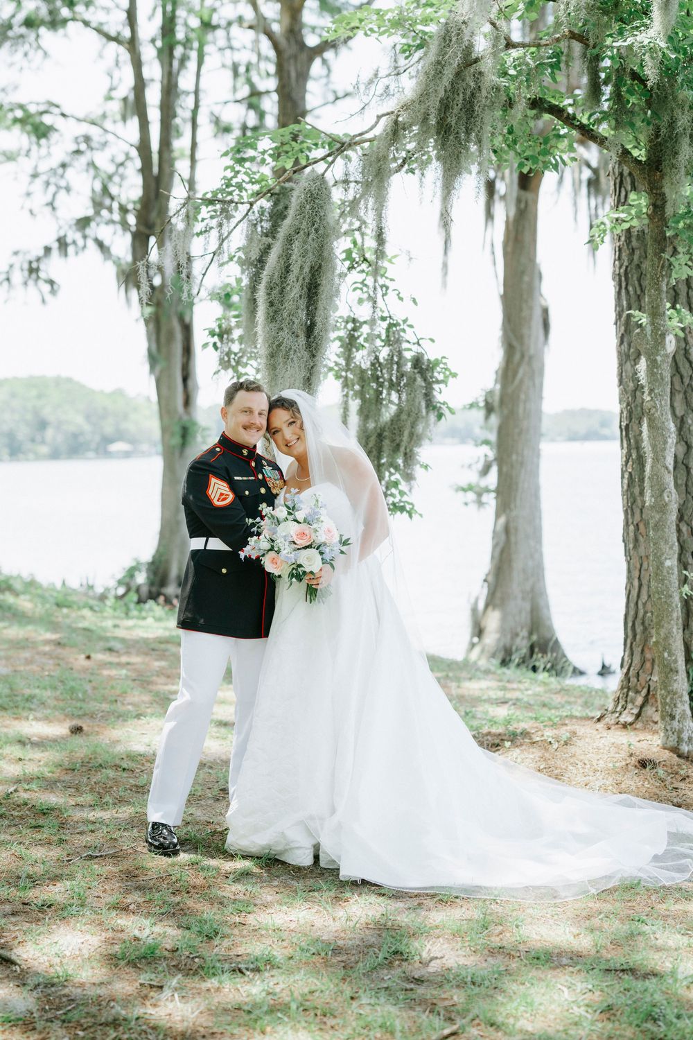 Marine groom in dress uniform stands with bride in white gown holding floral bouquet under moss-draped trees by waterfront.