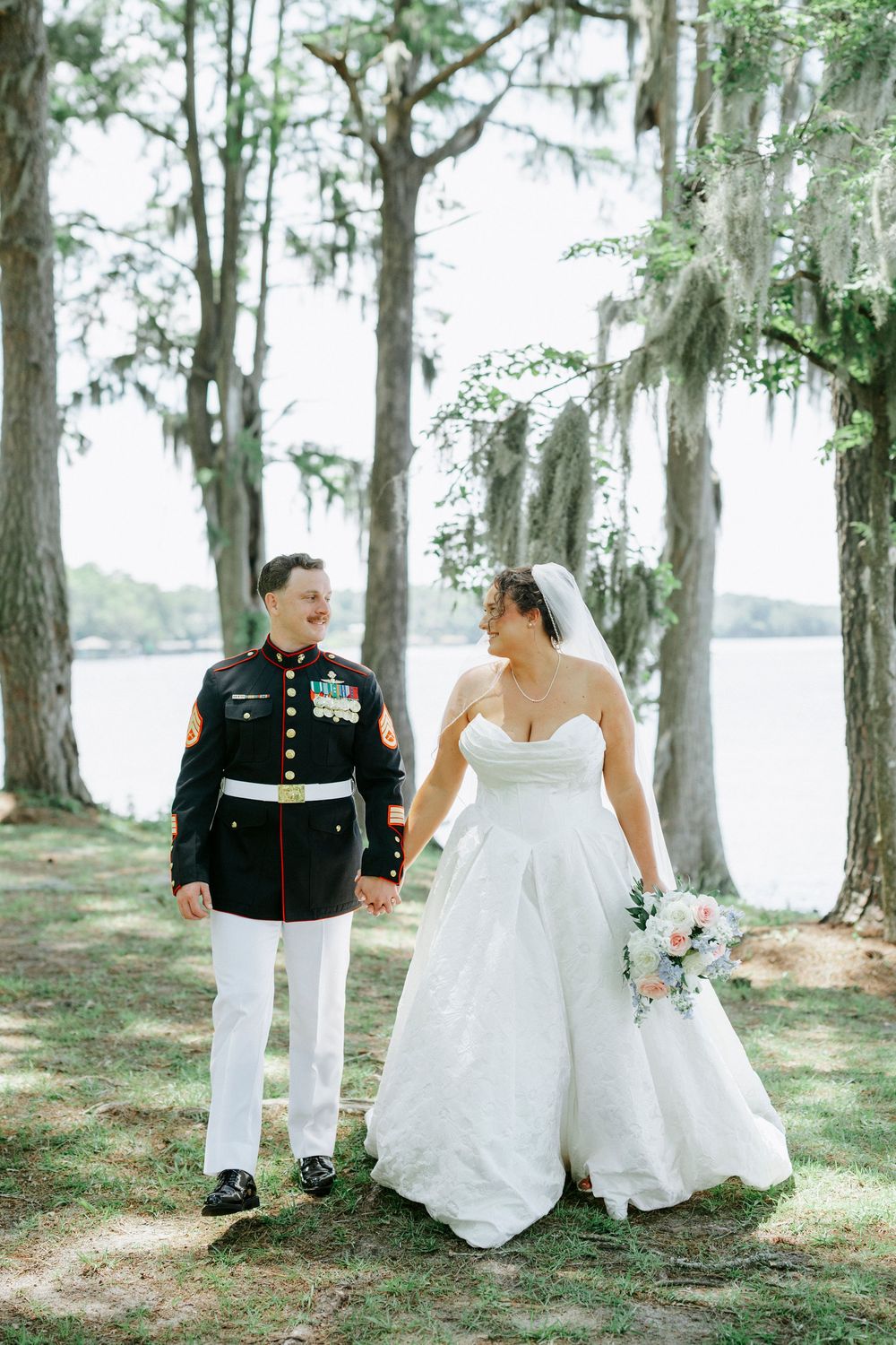 Marine groom in dress uniform holds hands with bride in white gown among moss-draped trees by waterfront.