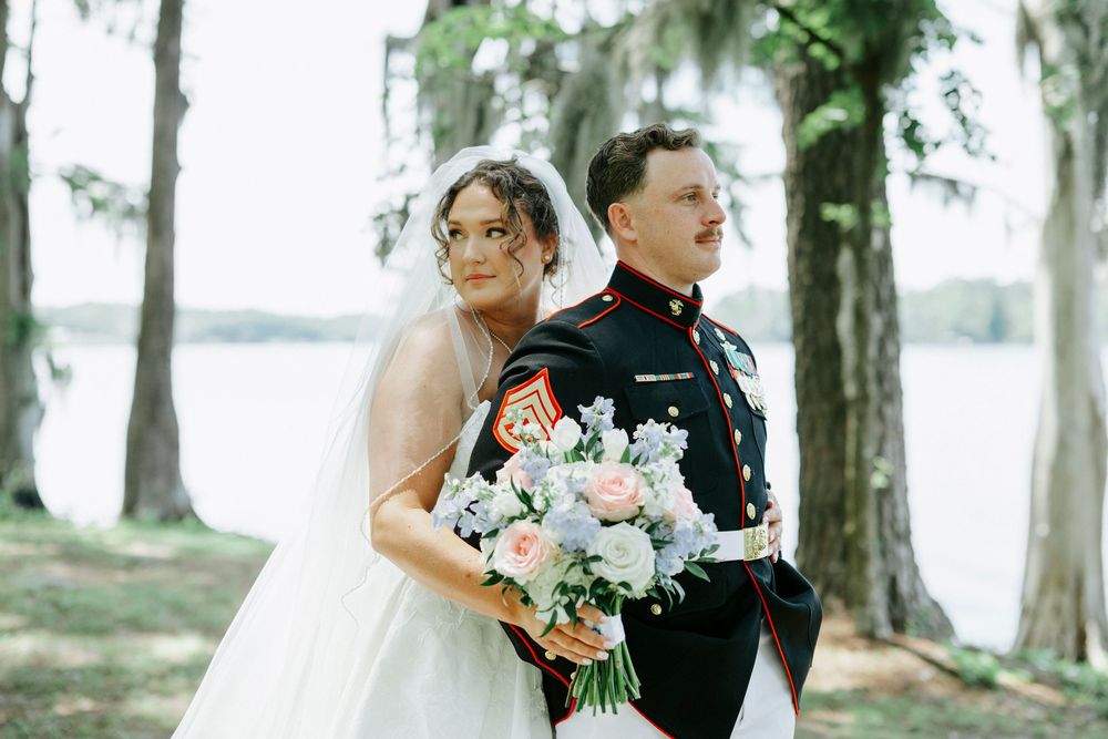 Bride in white gown and groom in Marine dress uniform stand back-to-back outdoors near a lake.