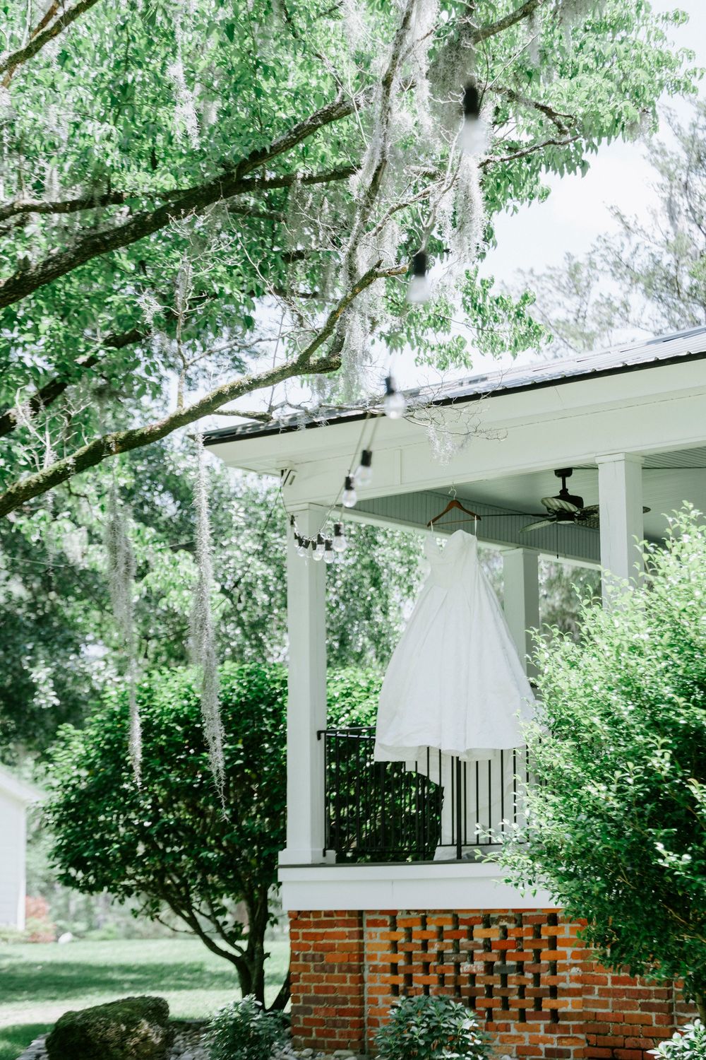 White wedding dress hanging on porch of brick venue surrounded by lush green trees.