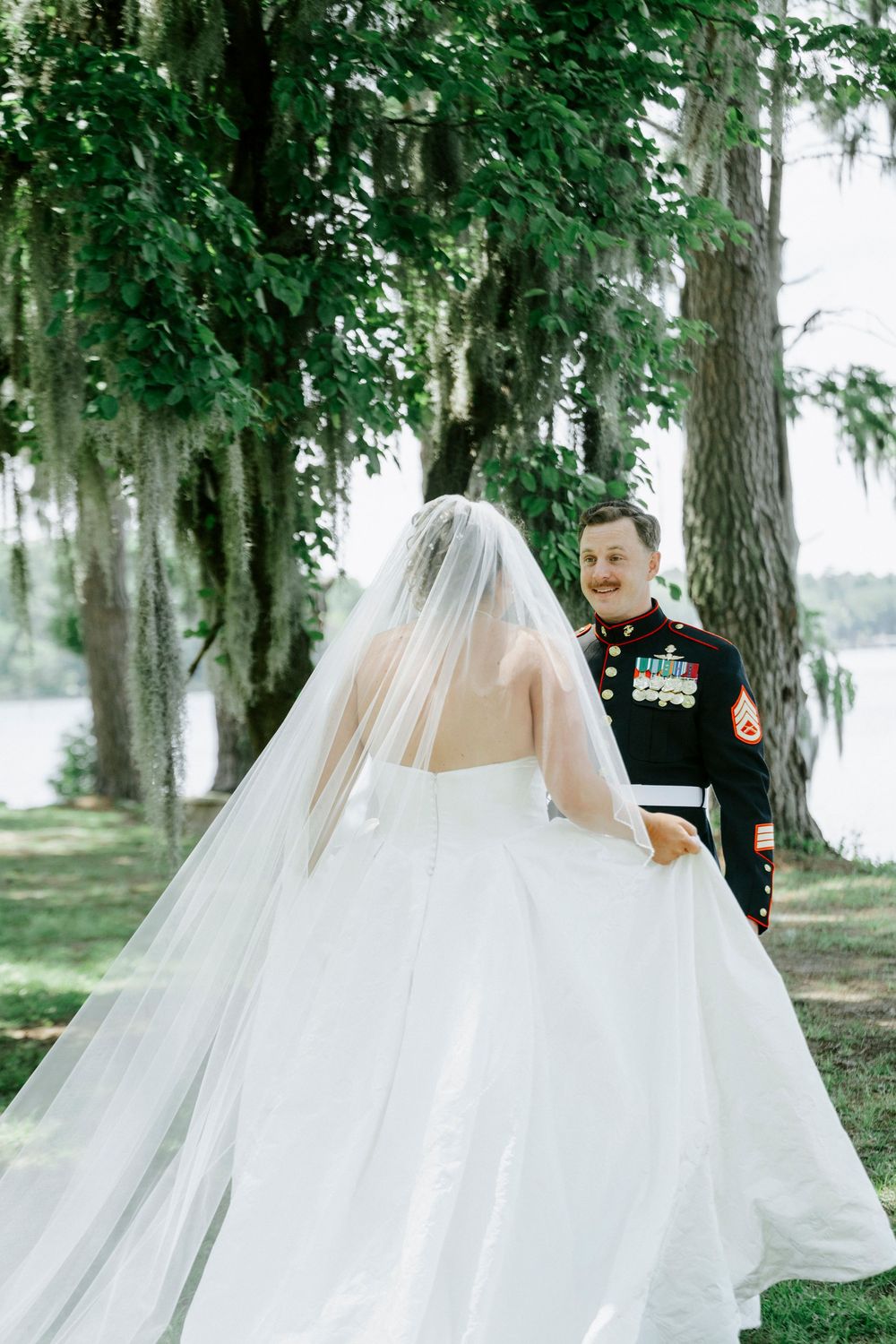 Marine groom in dress uniform smiles at bride in flowing white gown and veil outdoors near trees.