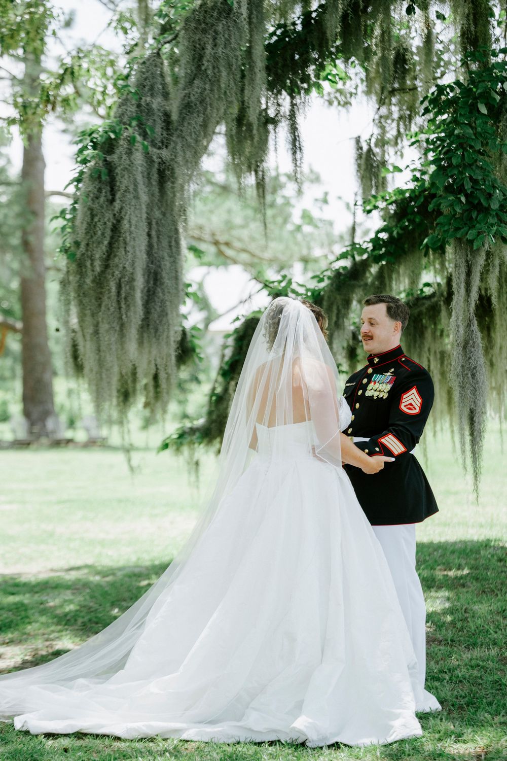 Bride in white gown and veil stands with Marine groom in dress uniform outdoors under Spanish moss-draped trees.