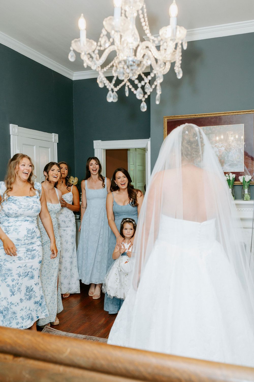 Bridesmaids in blue dresses react with joy as bride in white gown and veil reveals her look.