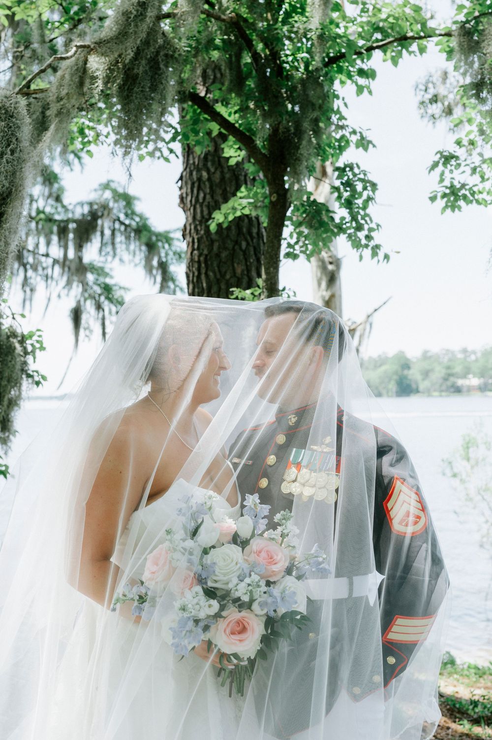 Military groom and bride sharing a kiss under her veil, holding a pastel bouquet outdoors near a lake.