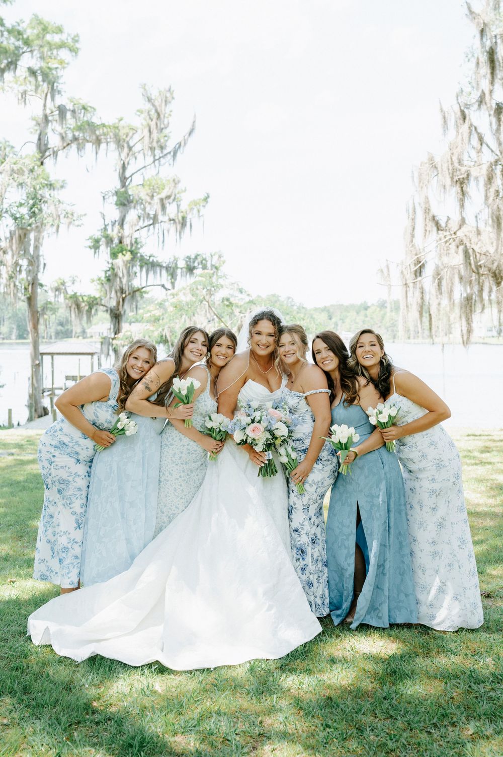 Bride in white gown poses with six bridesmaids in blue floral dresses holding bouquets outdoors near a lake.