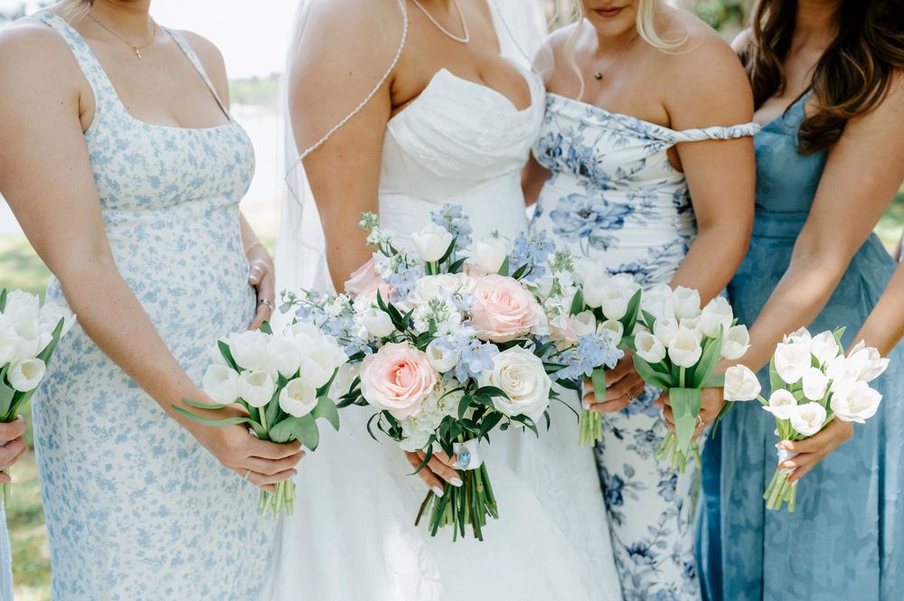 Bride in white gown holds pink and blue bouquet flanked by bridesmaids in floral blue dresses with white tulip bouquets.