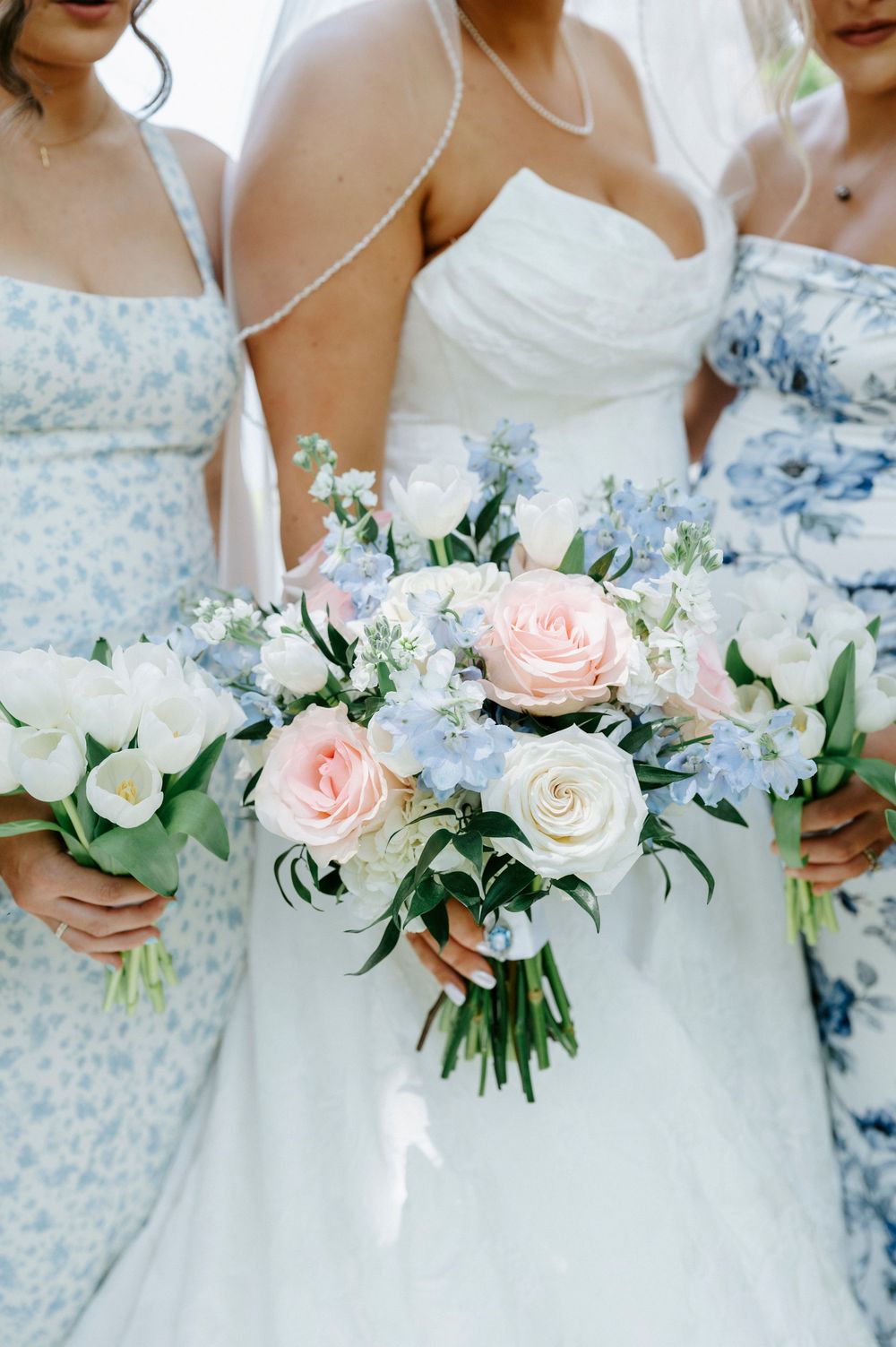 Bride and bridesmaids hold bouquets of pink roses, white flowers, and blue delphinium at wedding ceremony.