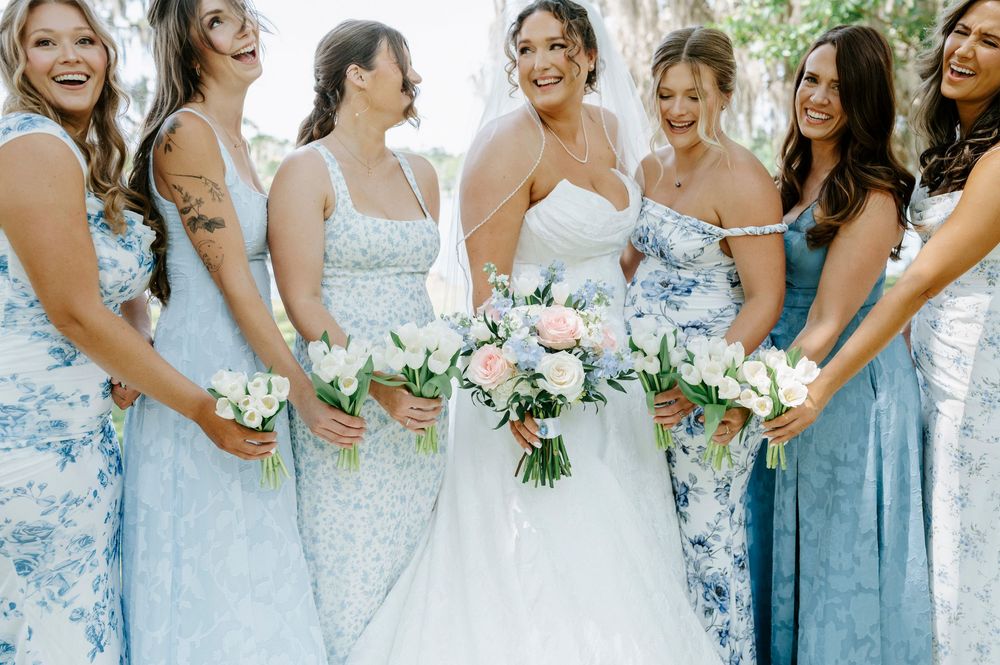 Bride and bridesmaids in light blue dresses laugh together while holding white and pink floral bouquets.