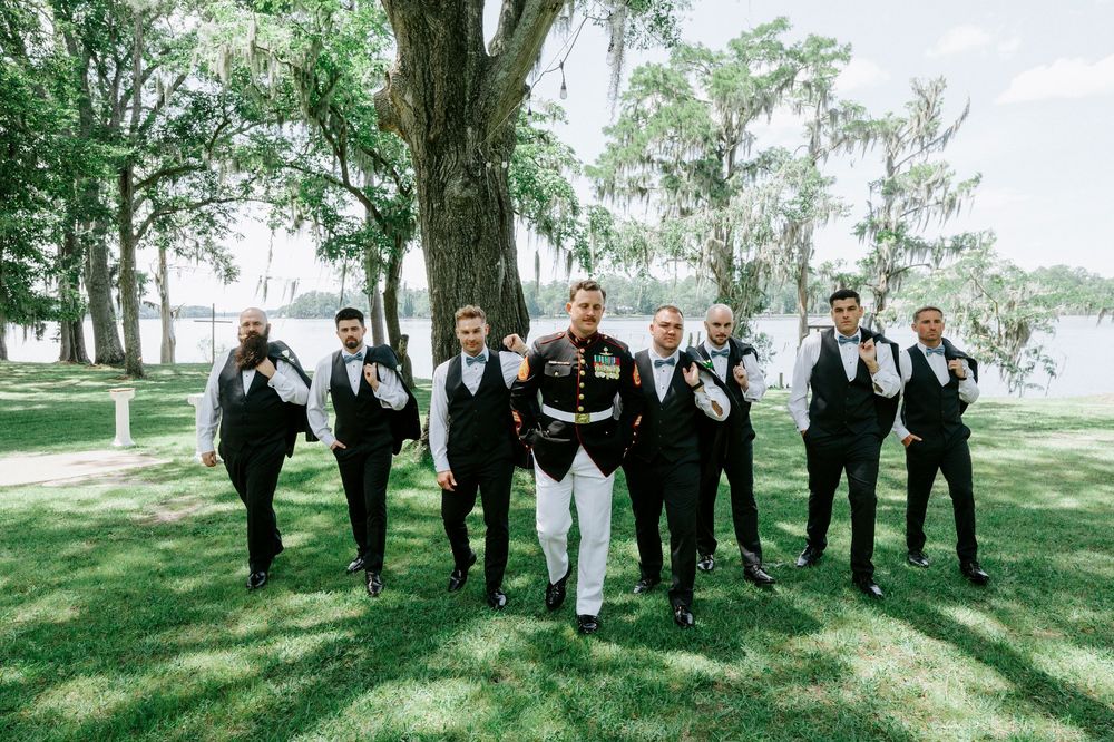 Marine groom in dress uniform walks with groomsmen in black suits on green lawn with moss-draped trees.