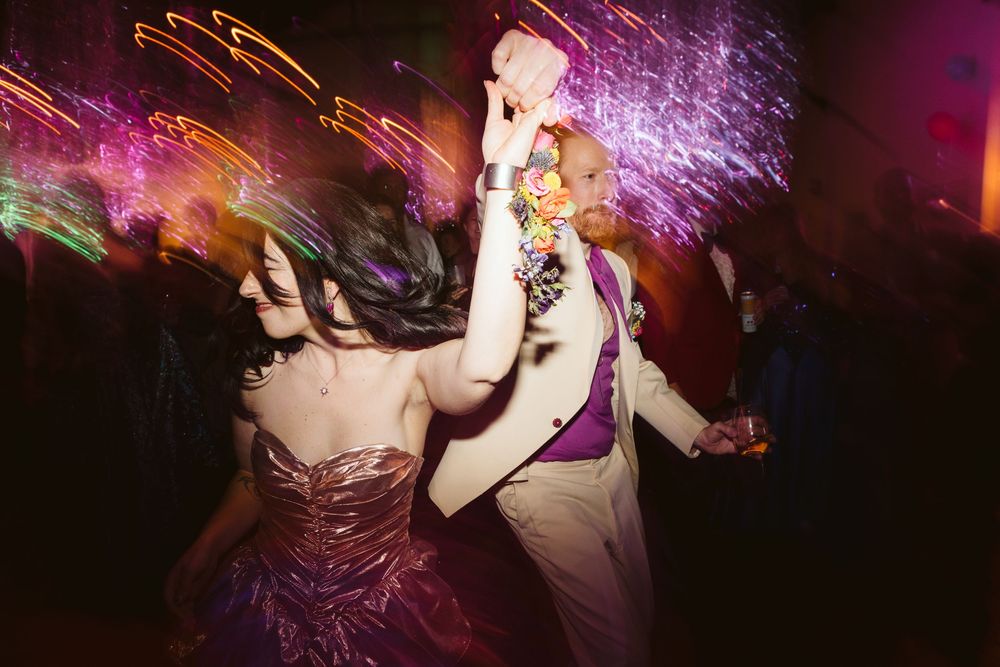 Couple dancing energetically at a vibrant wedding reception with colorful light streaks filling the dark dance floor.