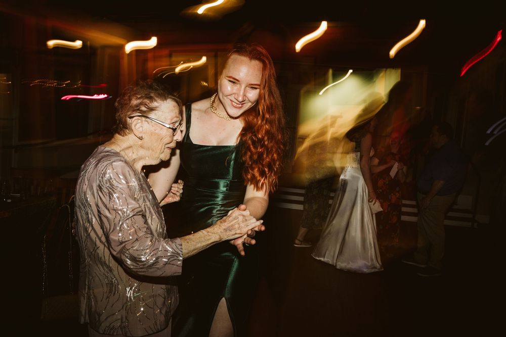 Young redhead woman in green dress dancing joyfully with elderly woman at a dimly lit party.