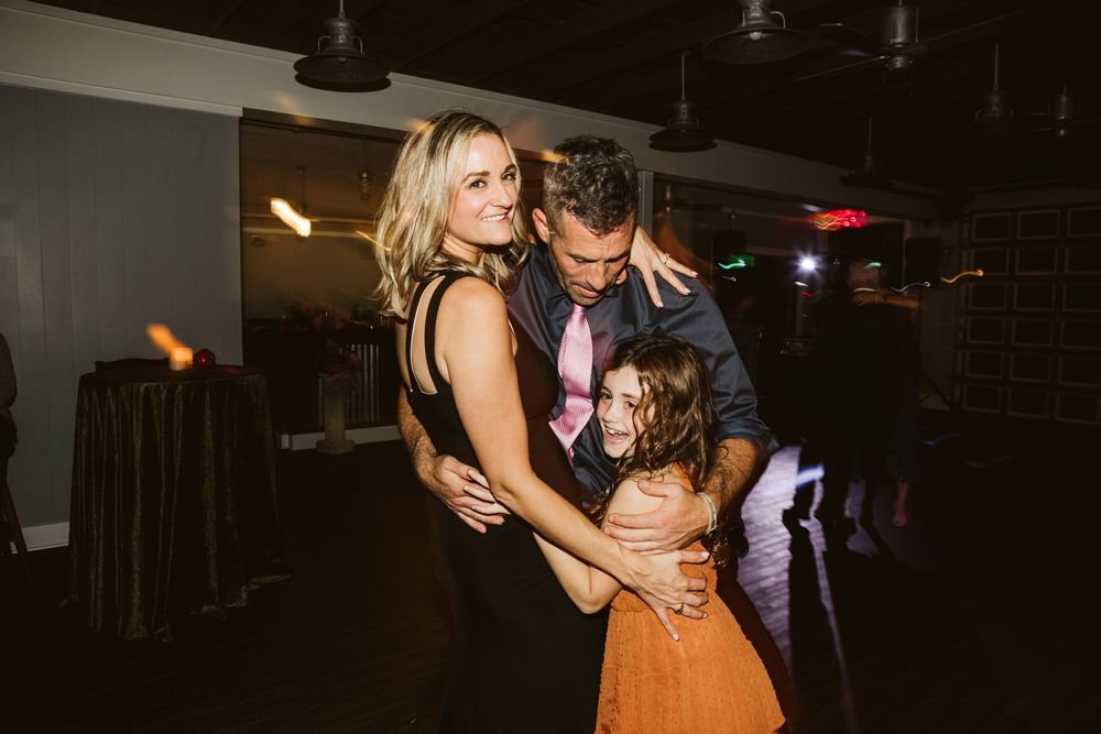 Family dancing together at a wedding reception, woman in black dress hugging man in suit and young girl in orange skirt.