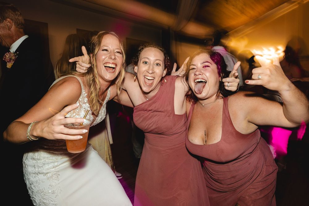 Three women celebrating at a wedding reception, laughing and posing together, bride in white gown holding a beer.