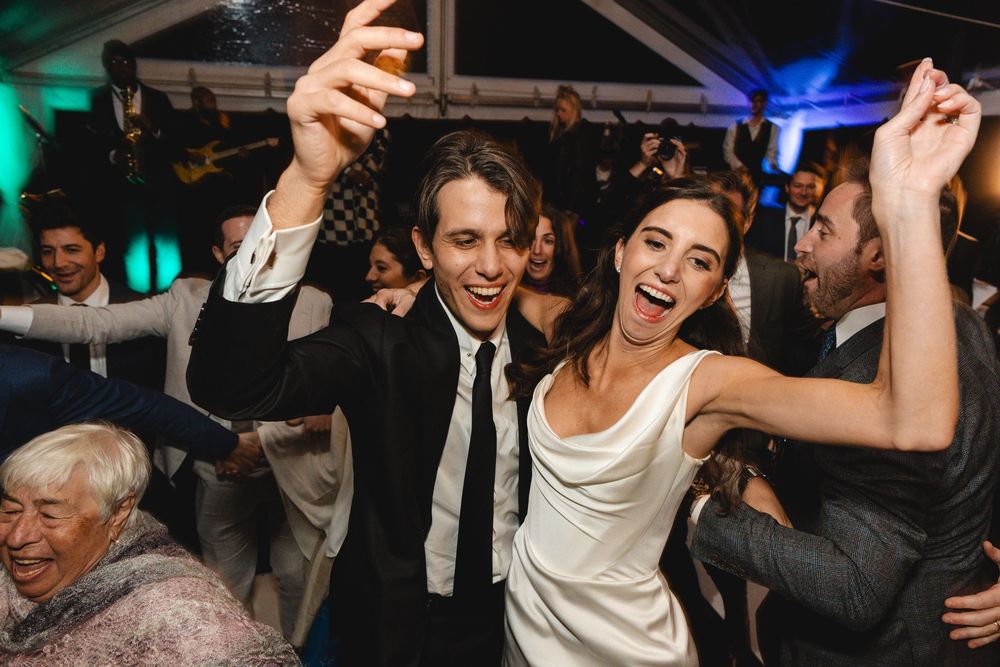 Joyful bride and groom dancing at wedding reception, raising arms in celebration among guests.