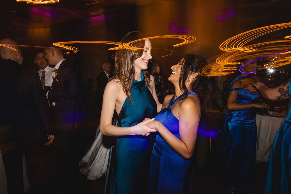 Two women in elegant blue gowns dancing joyfully at a formal event with swirling light trails.