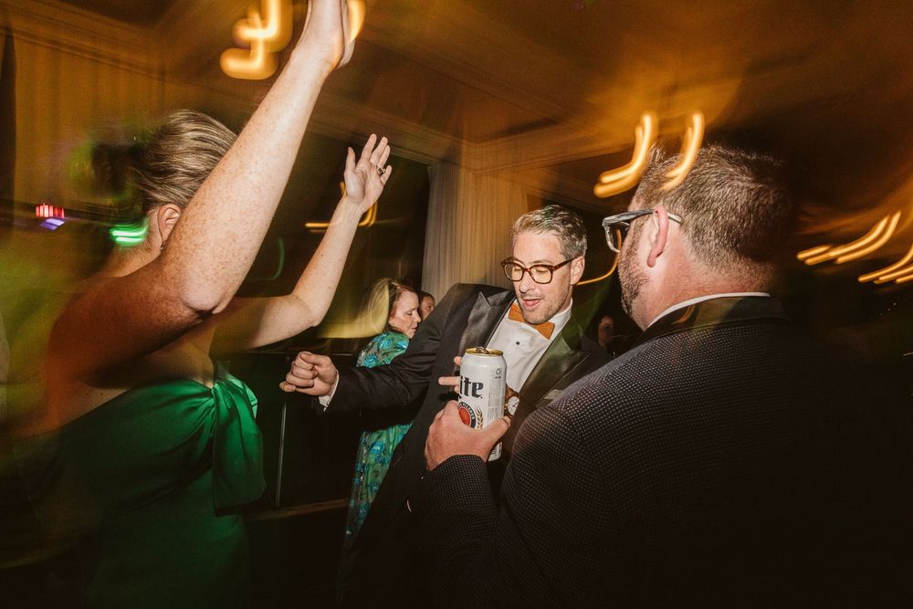 Guests dancing and celebrating at a wedding reception, holding drinks and raising hands in a dimly lit venue.