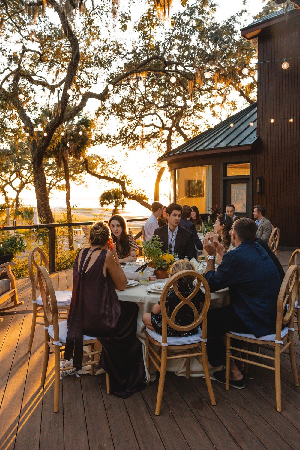 Guests dining al fresco on a wooden deck at sunset, surrounded by oak trees and string lights near a rustic building.
