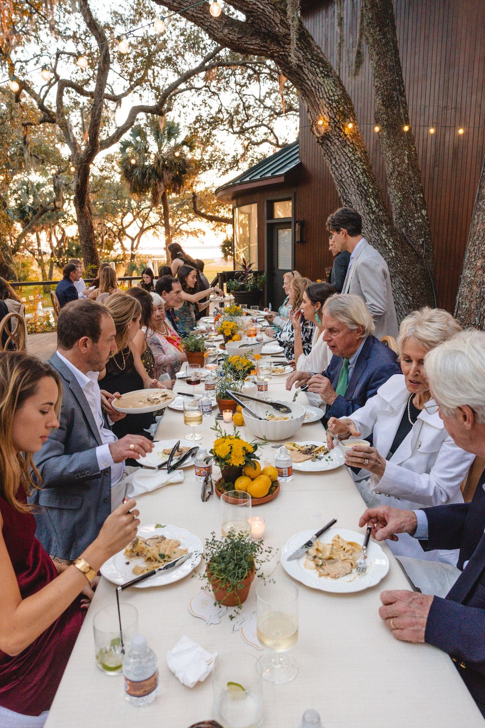 Guests dining at an elegant outdoor dinner party under oak trees with string lights at sunset.