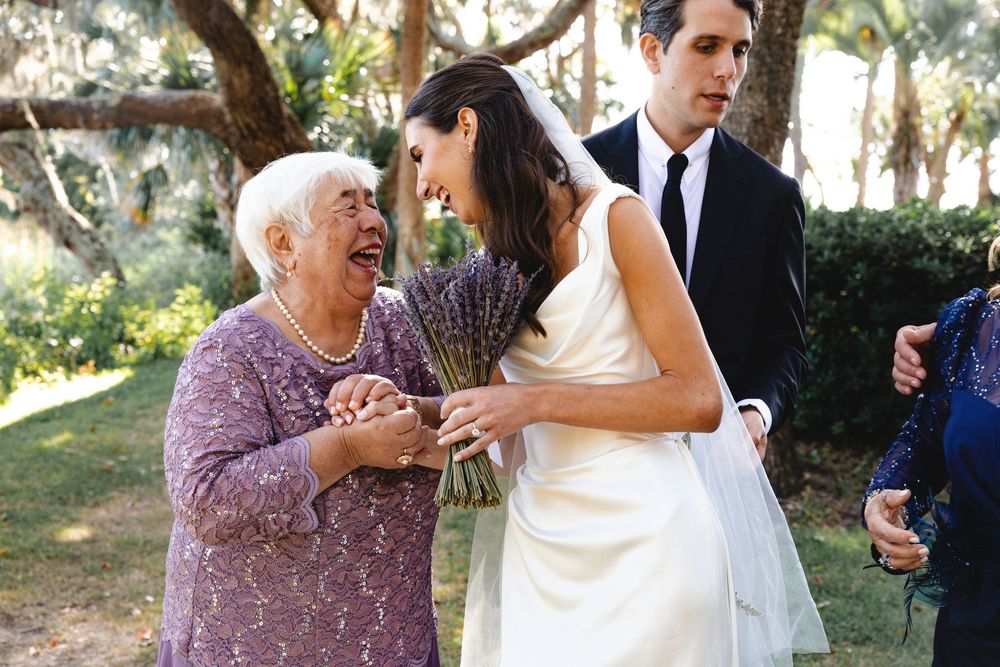 Elderly woman laughing joyfully with a bride holding lavender bouquet at an outdoor wedding celebration.