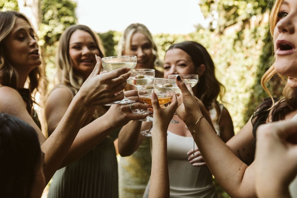 Group of women in elegant attire toasting with cocktail glasses at an outdoor celebration.