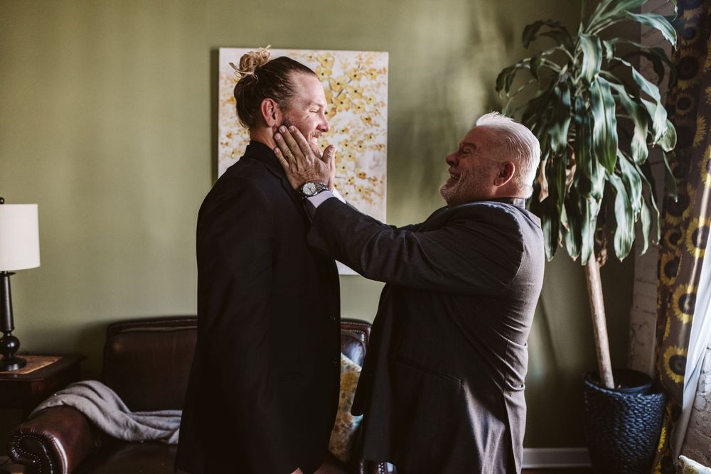Two men in suits share an emotional moment, one tenderly holding the other's face in a warmly decorated room.