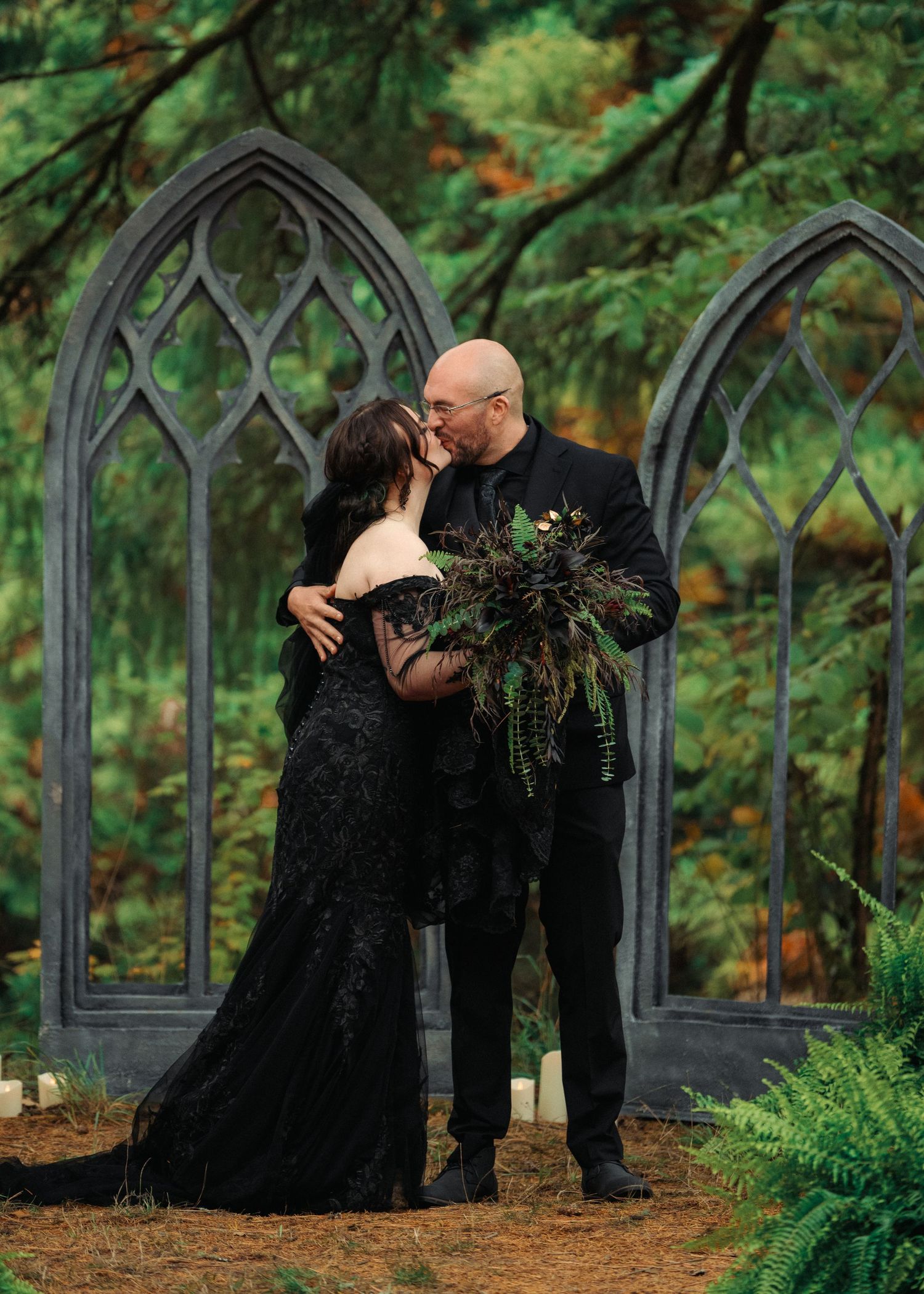 Couple sharing a romantic kiss in black wedding attire before gothic arch backdrop in a lush forest setting.