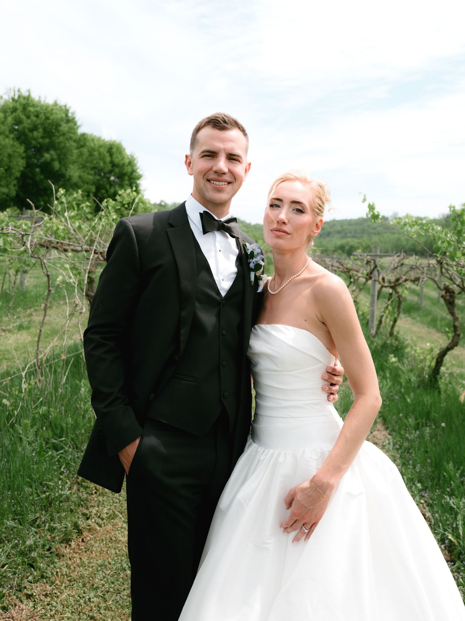 Black and white photo of elegant bride in strapless gown and groom in tuxedo posing outdoors at vineyard wedding.