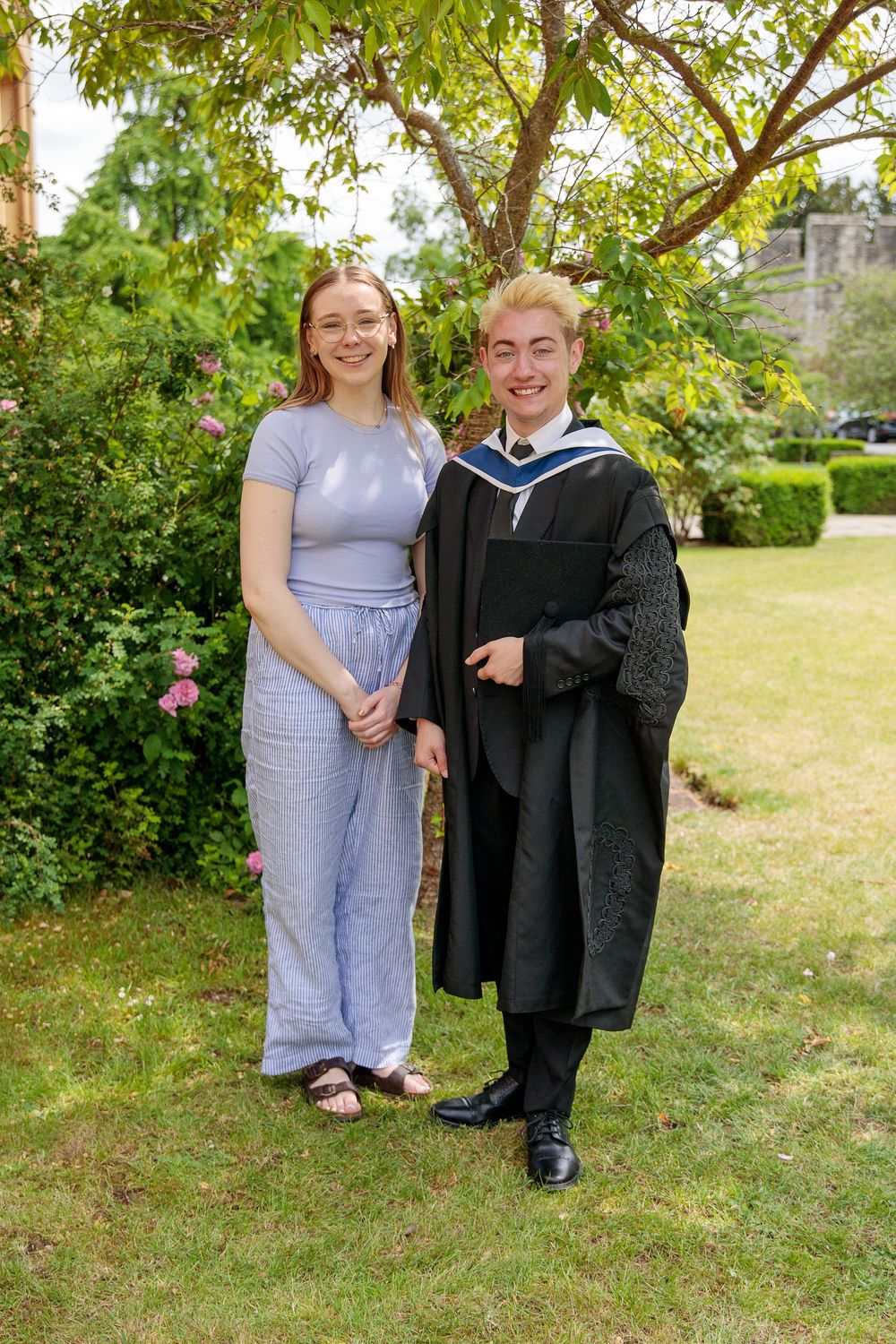 Outdoor graduation portraits with people posing on grass near flowering bushes.