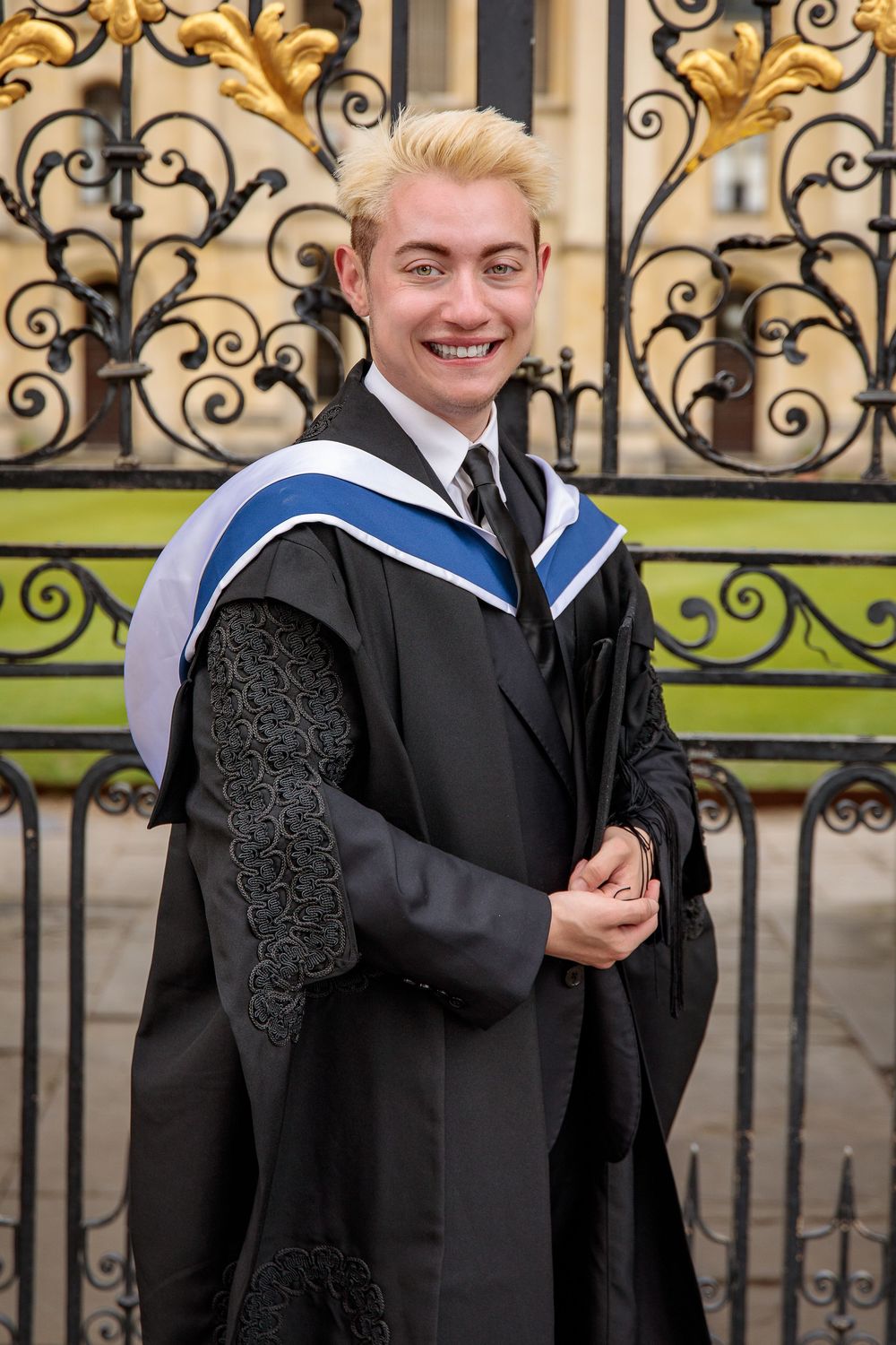 Graduate in academic dress posing by ornate gold and black iron gates.