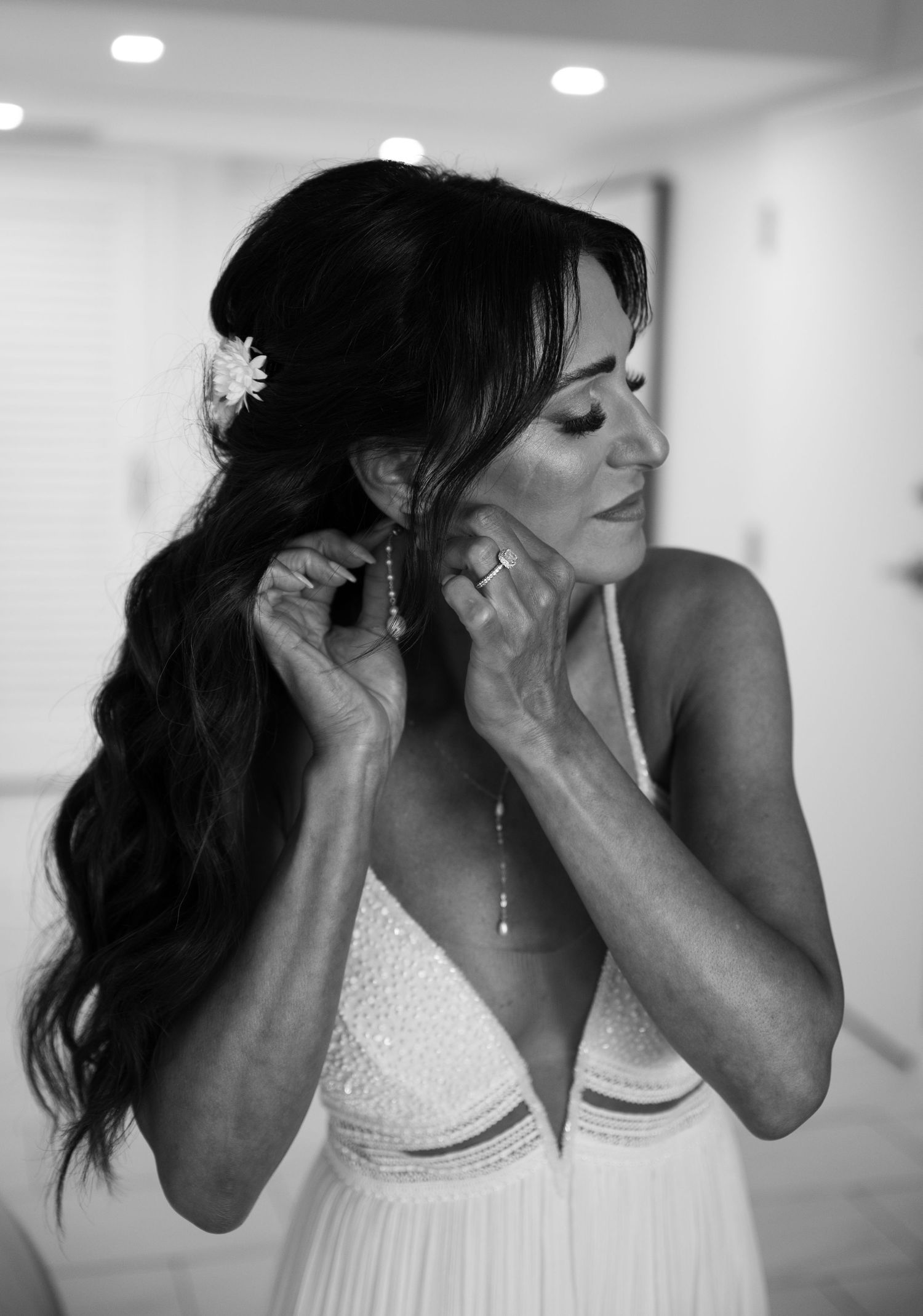 Bride in white gown adjusting earrings, long dark wavy hair with flower, black and white wedding photo.